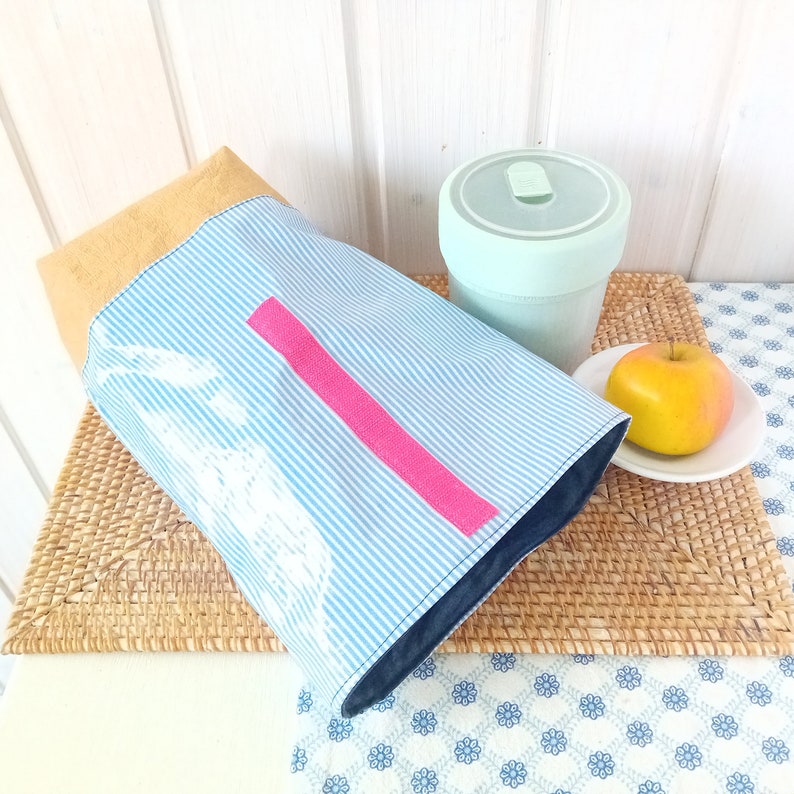 May include: A blue and white striped lunch bag with a pink stripe and a white design. The bag is sitting on a woven placemat next to a light green reusable cup and a yellow apple.