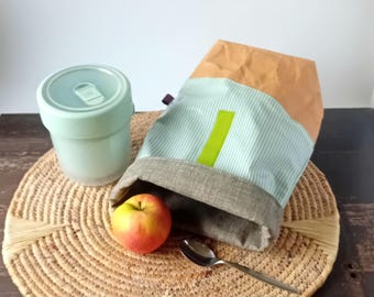 May include: A light green lidded food container, a tan and light green striped fabric lunch bag with a lime green vertical accent, a red and yellow apple, and a silver metal spoon. The items are arranged together on a round woven mat.