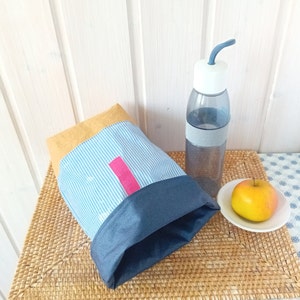 May include: A blue and white striped lunch bag with a yellow top and a pink zipper pull. The bag is sitting on a woven mat next to a clear water bottle with a blue lid and a blue straw. There is a yellow apple on a white plate.