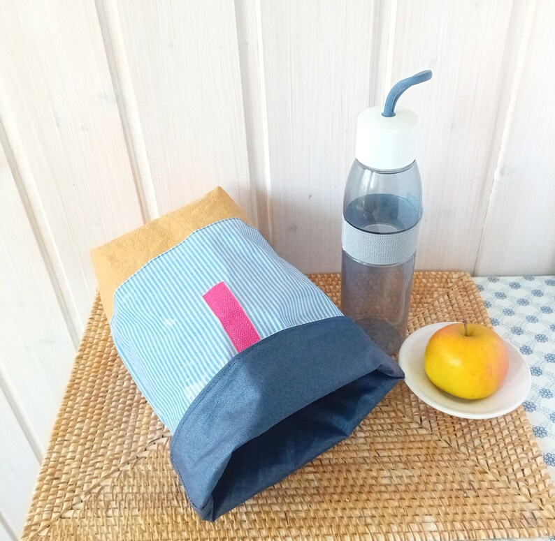 May include: A blue and white striped lunch bag with a yellow top and a pink zipper pull. The bag is sitting on a woven mat next to a clear water bottle with a blue lid and a blue straw. There is a yellow apple on a white plate.