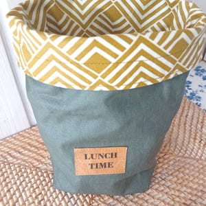 Könnte beinhalten: Eine grüne Stoff-Lunch-Tasche mit einem gelben und weißen geometrischen Muster am oberen Rand. Die Tasche hat einen braunen Leder-Patch mit dem Text "LUNCH TIME" darauf.