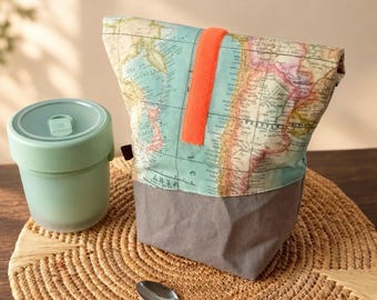 May include: A fabric lunch bag featuring a vintage world map print in shades of blue, green, and tan, accented by a bright orange vertical strap. Beside it sits a light green lidded container and a small silver metal spoon.