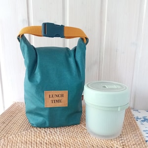 Könnte beinhalten: Türkisfarbene Lunchtasche mit senfgelbem Griff und Schnalle, neben einem hellgrünen Lebensmittelbehälter. Die Lunchtasche hat ein rechteckiges Etikett mit der Aufschrift "LUNCH TIME".