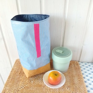May include: A blue and white striped fabric lunch bag with a pink handle and a brown bottom. The bag is sitting on a woven placemat next to a green reusable container and a red apple.