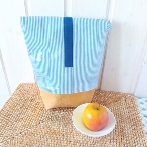 May include: A blue and white striped fabric bag with a blue strap and a brown bottom. The bag is sitting on a woven placemat next to a red apple on a white saucer.