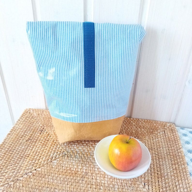 May include: A blue and white striped fabric bag with a blue strap and a brown bottom. The bag is sitting on a woven placemat next to a red apple on a white saucer.