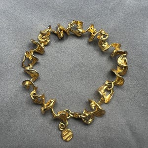 May include: A gold-toned bracelet with an abstract, swirling design. The bracelet features a series of interconnected, organic shapes. A small, circular charm with text hangs near the clasp. The bracelet is displayed on a gray surface.