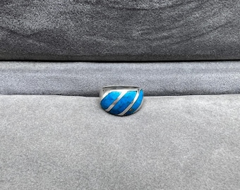 Puede incluir: Un anillo de plata con una banda ancha que presenta una vibrante incrustación turquesa con rayas plateadas diagonales. El anillo está sobre un fondo gris, mostrando su diseño y artesanía.