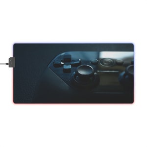 Könnte beinhalten: Eine schwarze Gaming-Mausmatte mit einem blauen und roten LED-Lichtrahmen. Ein schwarzer Videospiel-Controller liegt auf der Matte.