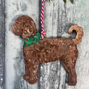 May include: A brown wooden Christmas ornament shaped like a dog wearing a green and red holly garland. The ornament is hanging from a red and white string.