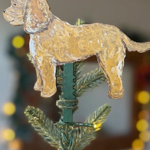May include: A golden-coloured dog ornament with a textured, painted finish, attached to a dark green clip. The dog is depicted in a standing profile position with floppy ears and a long tail, designed to be secured onto a branch of a tree.