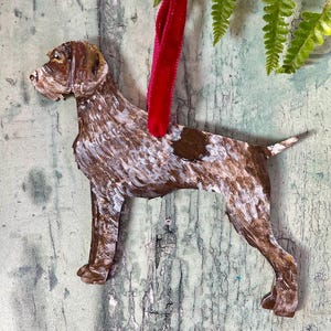 May include: Wooden dog ornament in a standing pose, painted with brown, white, and tan hues. The dog has a dark brown spot on its side and is suspended by a red velvet ribbon. Fern fronds are visible in the background.
