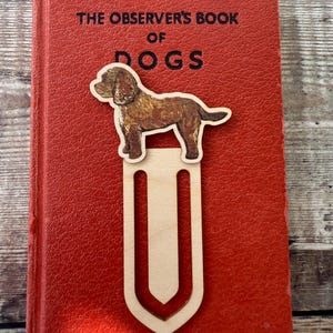 May include: A light-colored wooden bookmark shaped like a large paperclip, featuring a brown dog illustration attached to the top. The bookmark is resting on a red book titled The Observer's Book of Dogs, with black text printed on the cover.