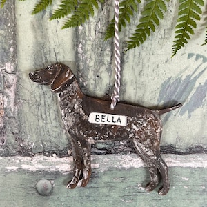 May include: A metal dog ornament with a rustic finish, shaped like a German Shorthaired Pointer. The ornament has a white tag with the name "Bella" written on it.