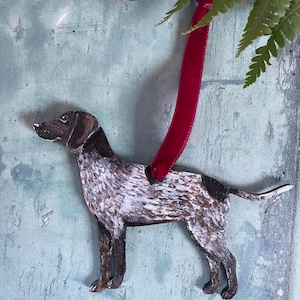 May include: A wooden ornament of a brown and white spotted dog with a red velvet ribbon attached. The dog is standing on a light blue background.