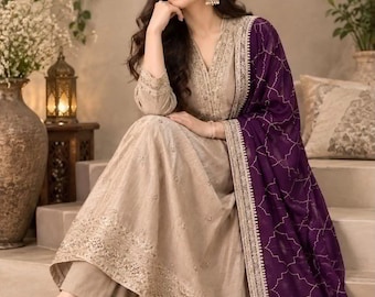 May include: A beige long-sleeved tunic dress with intricate gold embroidery along the neckline and hem, paired with matching beige trousers and a deep purple dupatta featuring a delicate white geometric pattern and a wide gold border.