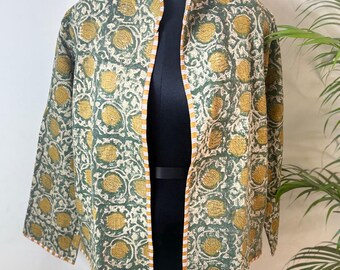 May include: A green and yellow floral print jacket with a mandarin collar. The jacket is open in the front and has three-quarter sleeves. The trim is a striped orange and white. The jacket is displayed on a black mannequin.