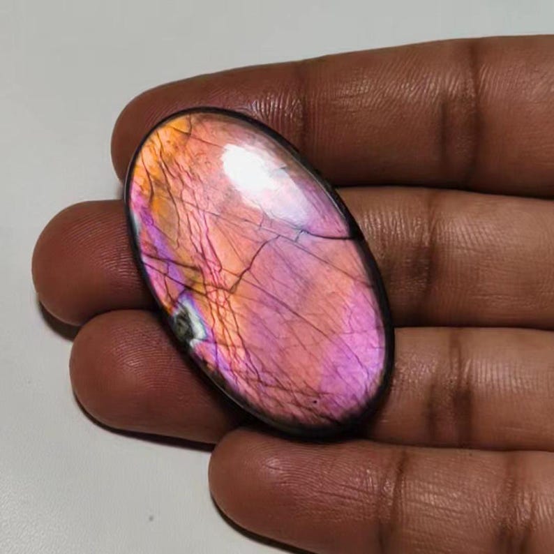 May include: Oval-shaped labradorite gemstone with iridescent pink, purple, and orange hues. The stone has a smooth, polished surface and is set in a silver bezel.