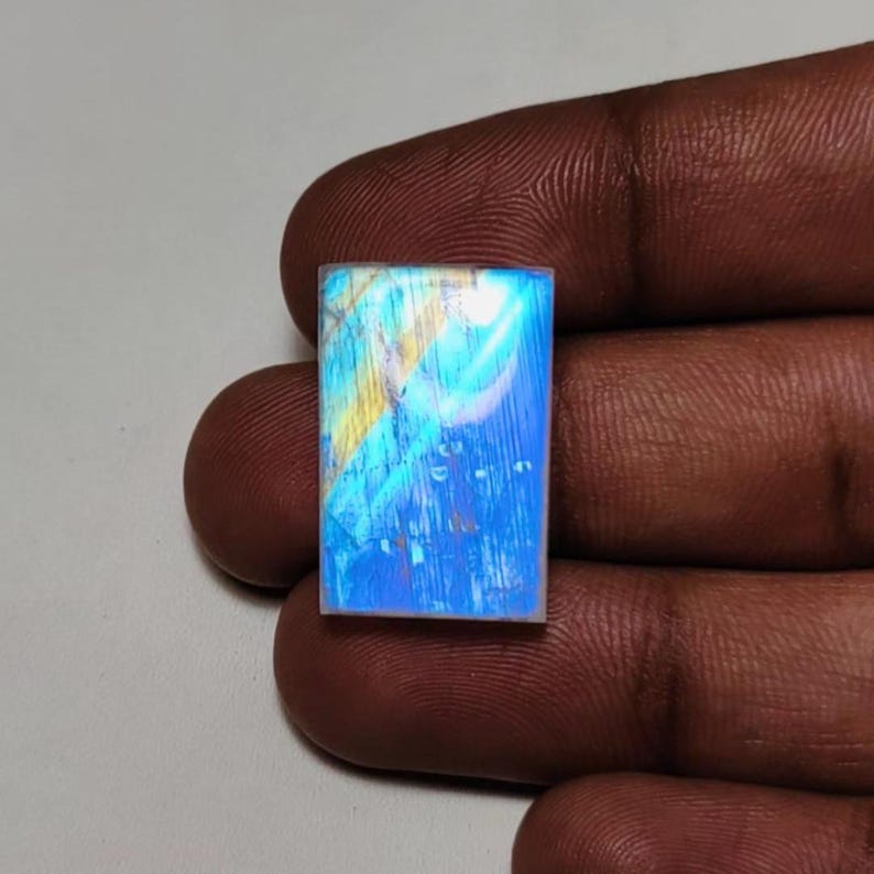 May include: A rectangular moonstone gemstone featuring a smooth, flat surface with a prominent blue adularescence and subtle yellow streaks visible across the translucent, polished stone face, showcasing a distinct iridescent glow.