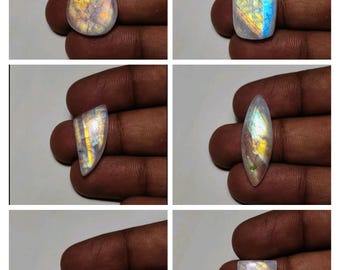 May include: Six polished moonstone gemstones in various shapes, including teardrop, rectangular, and oval. The stones display iridescent colors of blue, yellow, and orange, set against a translucent white base. Each stone is held in a hand.