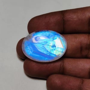 May include: An oval-shaped moonstone cabochon featuring a translucent, milky white base with a vibrant, glowing blue adularescence that shifts across the surface. The gemstone has a smooth, polished, domed top and a flat bottom.