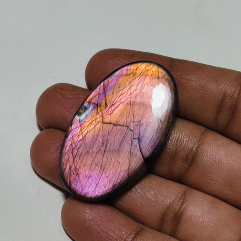 May include: Oval-shaped, iridescent labradorite gemstone with a pink, orange, and blue color scheme. The stone has a smooth, polished surface and a dark gray border.