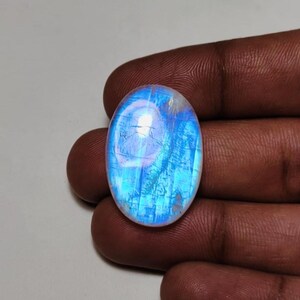 May include: An oval-shaped moonstone cabochon featuring a translucent, milky white base with a vibrant, glowing blue adularescence. The gemstone displays internal inclusions and a smooth, polished surface, reflecting light with a distinct blue sheen.