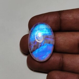 May include: An oval-shaped moonstone gemstone with a blue and white iridescent glow. The stone has a smooth, polished surface and is held by a hand. The gemstone's colors shift with the light, creating a captivating display.