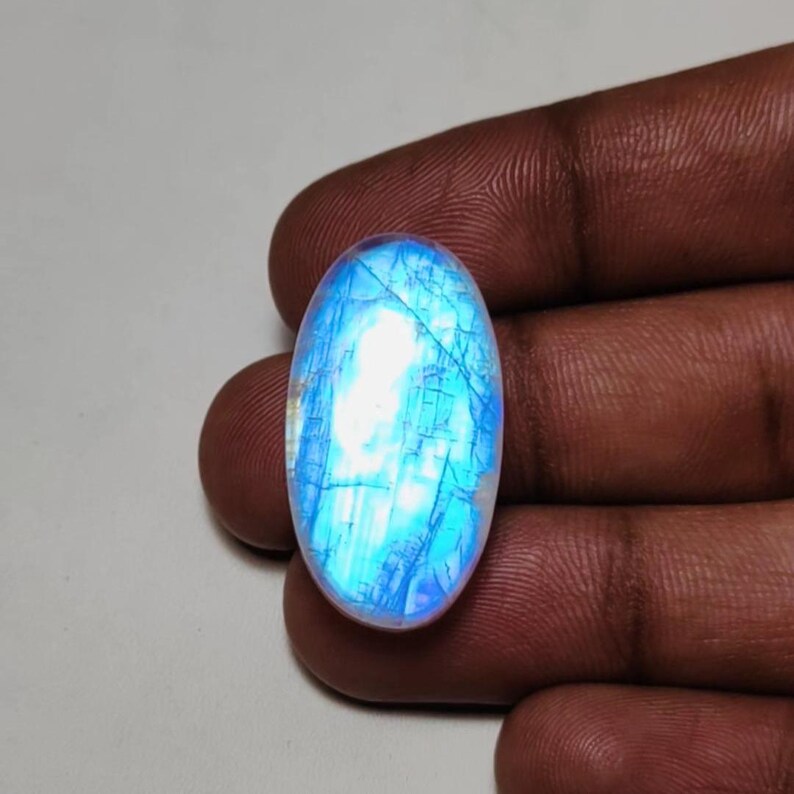 May include: Oval-shaped, iridescent moonstone gemstone. The stone displays a captivating play of light, with a predominantly blue and white color palette. The surface features delicate internal structures, enhancing its natural beauty. The gemstone is held between fingers.