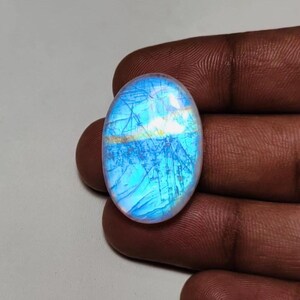 May include: Oval-shaped, iridescent gemstone with a blue and white color scheme. The stone has a network of fine lines across its surface, creating a unique pattern. The gemstone is held between fingers.
