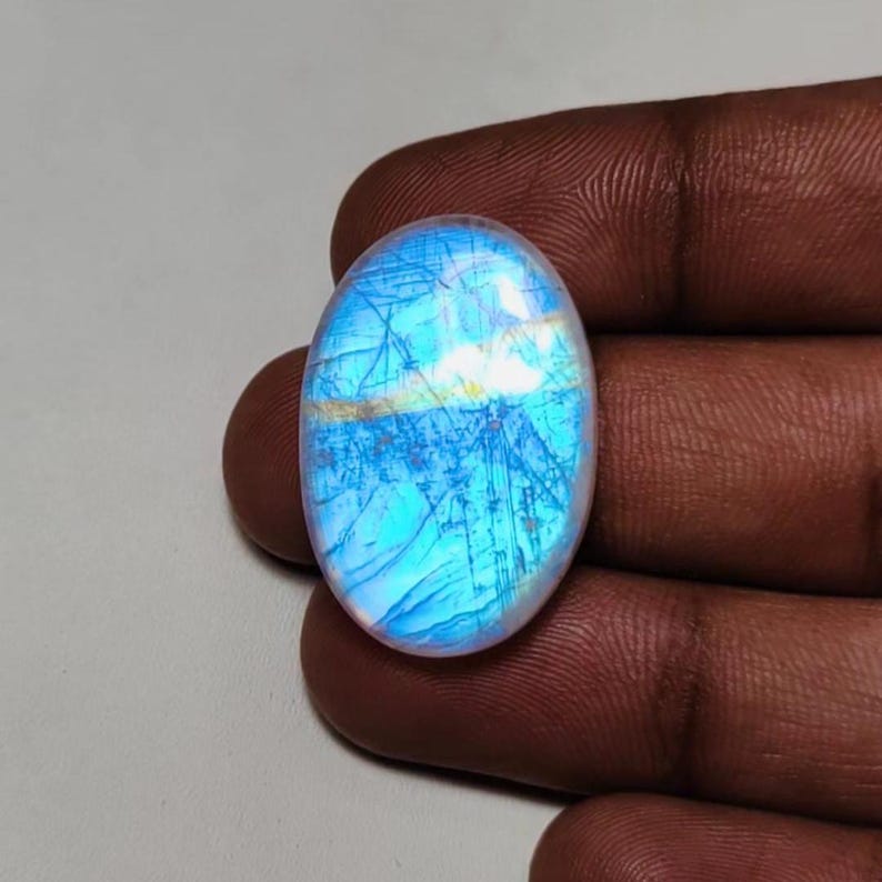 May include: Oval-shaped, iridescent gemstone with a blue and white color scheme. The stone has a network of fine lines across its surface, creating a unique pattern. The gemstone is held between fingers.