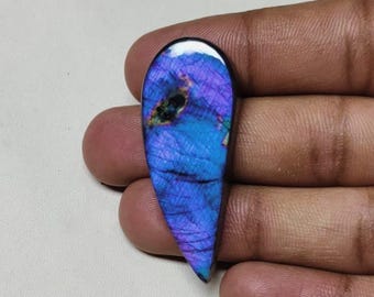 May include: A teardrop-shaped labradorite gemstone with a polished surface. The stone displays a vibrant play of color, with hues of blue, purple, and hints of green. The gemstone is held in a hand, showcasing its size and shape.