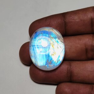 May include: Oval-shaped, iridescent moonstone gemstone. The stone displays a play of colors, with blues, whites, and hints of yellow. The polished surface reflects light, creating a shimmering effect. The gemstone is held in a hand.