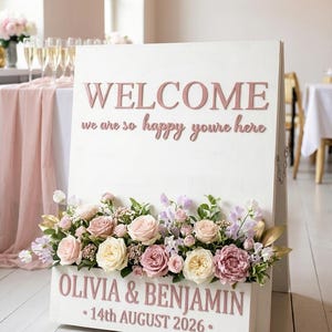May include: A white wooden welcome sign with pink lettering that reads "WELCOME, we are so happy you're here". The sign is adorned with a floral arrangement of pink and cream roses, and the names "OLIVIA & BENJAMIN" and the date "14th AUGUST 2026" are also displayed.