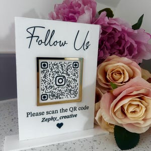 Könnte beinhalten: Ein weißes, rechteckiges Acrylschild mit dem schwarzen Text Follow Us oben, einem mittigen, goldgerahmten Instagram-QR-Code und dem Text Please scan the QR code Zephy_creative mit einem kleinen schwarzen Herzsymbol unten.