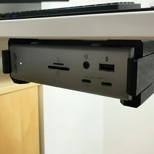 May include: A silver CalDigit docking station mounted under a white desk. The device has multiple ports, including USB, SD card reader, and audio jacks. The black mounting bracket is visible, and the CalDigit logo is on the side.