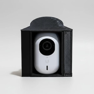 May include: A white security camera with a black lens and indicator light is enclosed in a black, rectangular housing. The housing has a curved top, similar to a small building. The camera is centrally positioned within the frame.
