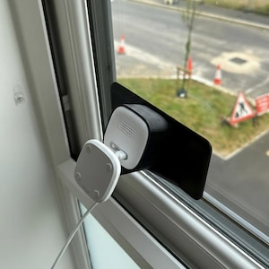 May include: A white and black security camera system mounted on a window. The camera has a square, rounded design and is connected to a white square power adapter with a white cable. The camera is attached to a black bracket.