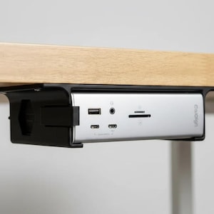 May include: A silver CalDigit docking station mounted in a black plastic bracket under a light wood surface. The front panel features a USB-A port, a 3.5mm audio jack, an SD card slot, and two USB-C ports with small icons printed below each port.