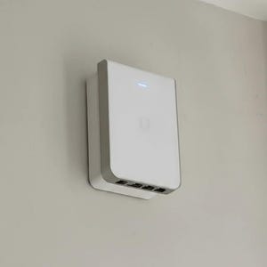 May include: A white, rectangular device with rounded corners is mounted on a light grey wall. The device has a grey border and a small blue light at the top. The bottom has several black ports. The letter "U" is visible on the front.