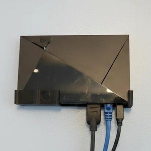 May include: A black rectangular media streaming device mounted in a black plastic wall bracket. The device features a geometric angular design and is connected to a black HDMI cable, a blue Ethernet cable, and a black power cable at the bottom.