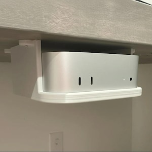 May include: A silver computer is mounted under a desk with a white bracket. The computer has two vertical slots and a small hole on the front. The bracket is white and has a curved front edge.