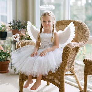 May include: A young child wearing a white angel costume, including a dress, wings, and a halo, sits on a wicker chair. The dress has a tulle skirt and a crochet bodice. A heart-shaped wand rests nearby. The scene is set indoors with natural light.