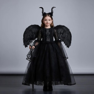 May include: A black costume featuring a long, layered dress, feathered wings, and a horned headband. The costume includes a staff with a decorative orb. The outfit is designed for a child and is suitable for Halloween or costume parties.
