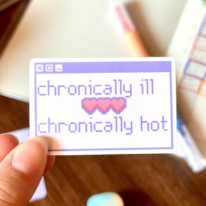 Puede incluir: Una pegatina blanca con un borde morado claro y el texto "chronically ill" y "chronically hot" en una fuente pixelada. La pegatina también presenta tres corazones rosas pixelados. La pegatina está sujeta por una mano.