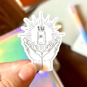 May include: A white sticker featuring a black and white illustration of a salt shaker held in cupped hands, with radiating lines behind it. The word "SALT" is printed on the shaker. The sticker is held in a hand against a blurred background.