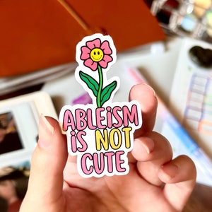 Könnte beinhalten: Ein Aufkleber mit einer rosa Blume und einem gelben Smiley-Gesicht. Der Text "ABLEISM IS NOT CUTE" ist in Rosa und Gelb dargestellt. Der Aufkleber wird in einer Hand gehalten. Der Hintergrund ist unscharf.