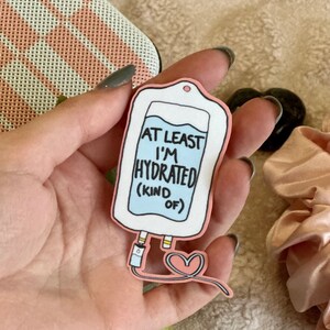 Może przedstawiać: Naklejka w kształcie worka do kroplówki z napisem "AT LEAST I'M HYDRATED (KIND OF)". Projekt zawiera jasnoniebieski płyn, różowe akcenty i serce.