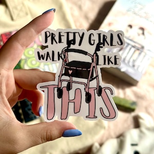 May include: A sticker with the text "PRETTY GIRLS WALK LIKE THIS" in black and pink. The sticker features a pink walker with a black seat. The sticker is held by a hand with blue nail polish.