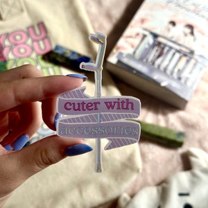 以下が含まれることがあります： 「cuter with accessories」の文字が入った、標識デザインのホログラフィックステッカー。本とトートバッグの前にステッカーが置かれています。